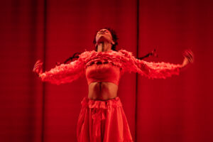 Artist Alessandra Azeviche mid dance performance. Wearing all red against a red curtained backdrop, arms outstretched, head towards the ceiling, the image shows a power and strenght in her movements. 