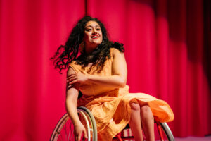 Artist Maryam Madani during her dance performance. She smiles brightly and her dress is a warm sunshine shade of yellow. We see movement in her hair swinging behind here. One hand on her whellchair the other gently towards her opposite arm.