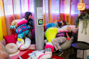 Visitors to Disrupt sitting amongst the soft sculptures created by artist Rion Duffy Murphy. The pieces are large, soft and squishy and are sitting amongst cushions, and blankets all brightly coloured. We see 2 people burrowed into the comfort of the space.