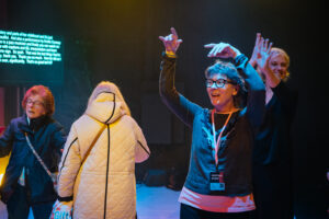 Audio Describer Mo Harte leads a touch tour on stage before a performance. She has her arms above her head and smiling, explaining what the next performances will be about. We see others on stage taking part in the touch tour.