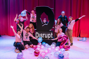 Disrupt Team all assemble around the Disrupt large foam logo. 9 people smiling and gesturing towards the camera, surrounded by ballons in blues and pinks.