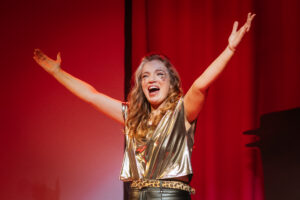 Artist Sarah Jane O'Regan closing out Disrupt's 2025 with her ISL Sign along Disco. Sarah Jane wears a gold shiny top and raises her arms to the sky in happiness and encouraging the audience to sing.