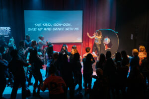Sarah jane O'Regan performing on stage. A room full of people all standing on the satge having fun and signing along.