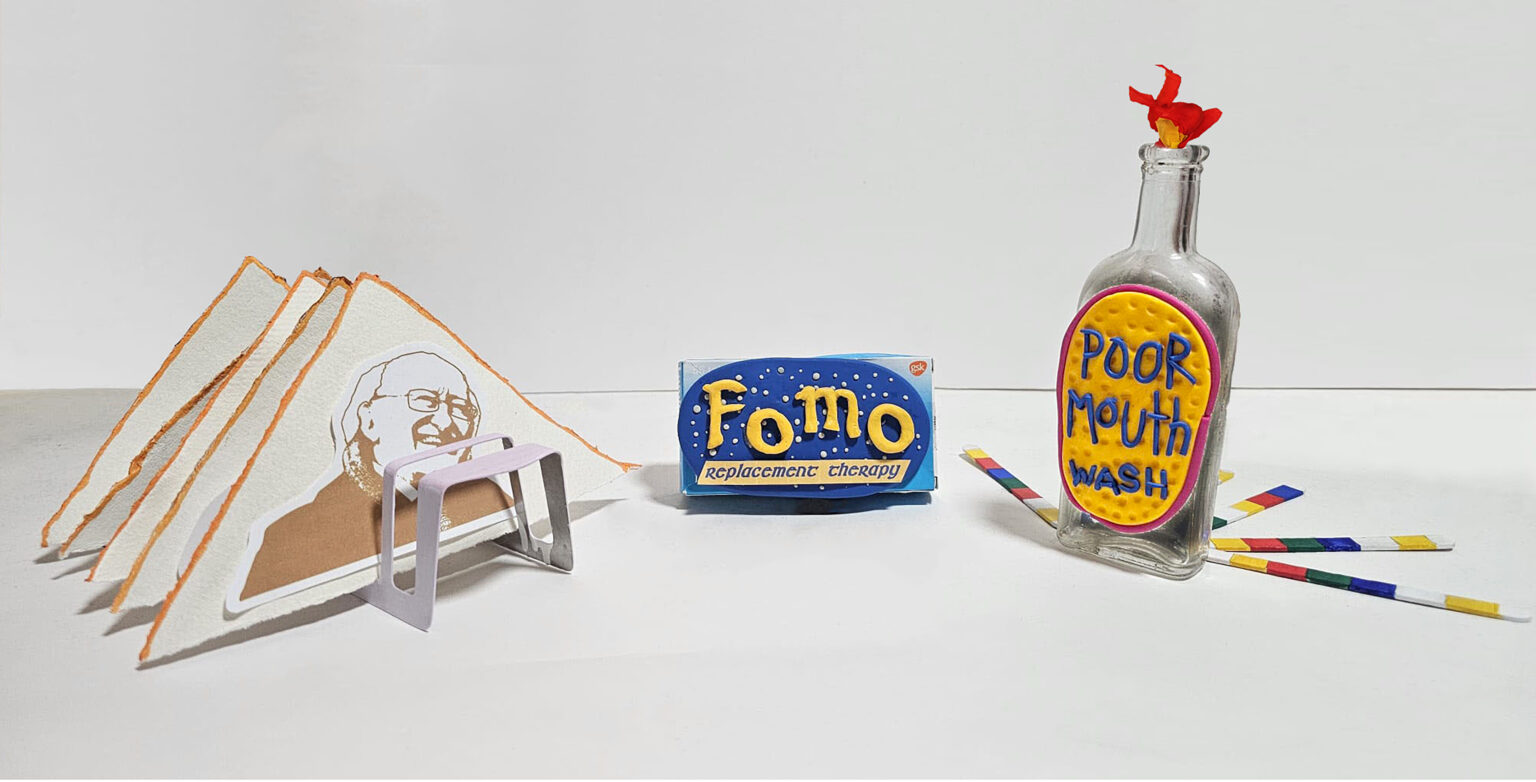 A photograph of a composition of an artwork made up of three elements sitting side-by-side. On a white background, the objects appear cartoony, handmade and humorous, but the images and text suggest a more serious message. To the left is a metal toast rack, painted light pink. Sitting in it are five triangular slices of bread made from rag paper. On the first slice is pasted a paper image of a smiling Michael D. Higgins, Ireland’s recent president. In the middle is a rectangular blue medication box with the letters GSK in the top corner. FOMO is spelled out in relief in big yellow letters across the front of the box, followed by the description Replacement Therapy. On the right is a tall, long-neck glass bottle with a crepe paper scarlet and yellow flame blossoming from the mouth of the bottle. It’s filled with ashes. It also has a 3D label, in bright primary colours, spelling Poor Mouth Wash. Thin sticks in primary colours splay out on the counter from the glass bottle, forming a bright semi-circle.