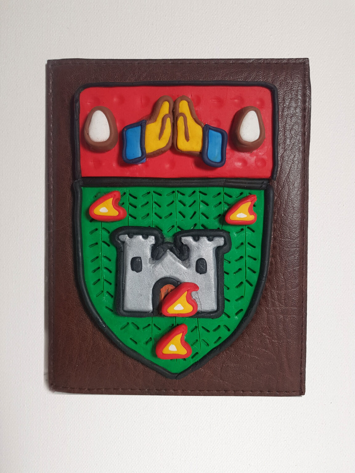 A photograph of small contemporary artwork. A heraldic crest is reimagined and remade in polymer clay, forming the shape of a shield. The symbolic imagery is reduced to simple graphics in bold block colours. In the top section, on a red background, are two hands clasped together in prayer, taking the style of an emoji. On either side are white tear-drop shapes. On the bottom, against a bright green background featuring a simple pattern incised into the polymer clay, is a silver-grey castle with a turret on either side and arched black windows. Flames surround the castle and lick at the wooden doors. The artwork is set onto brown faux-leather material, rectangular shaped with neat stitching around its edges.