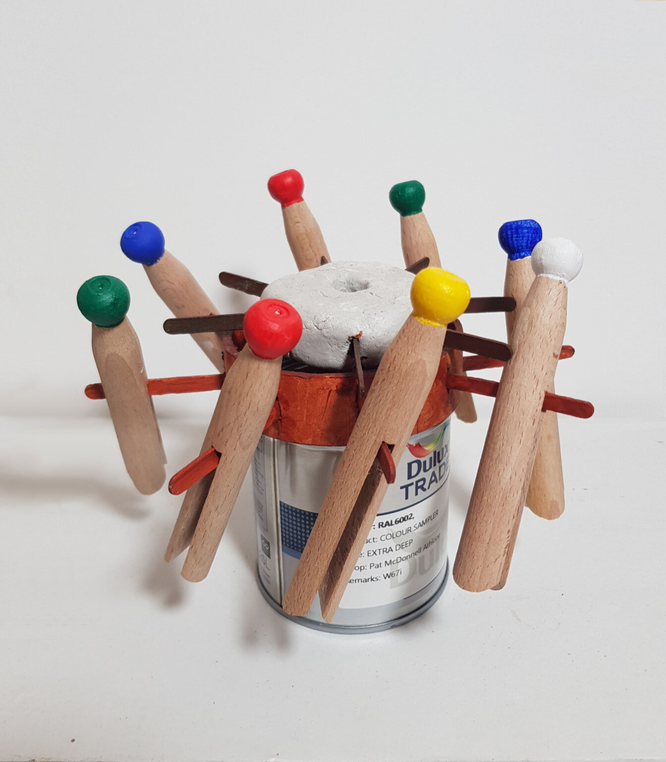 A photograph of a dynamic 3D sculptural artwork. Made of a variety of materials, it takes the form of a miniature capstan. A capstan is found on a sailing ship, typically made of iron, to help sailors secure heavy ropes. The artwork’s base is made from a sample-size tin of paint with the brand Dulux showing on the label. On top of the tin is a red lid with coffee stirrers protruding from it at regular intervals. Sitting astride the stirrers are eight wooden clothes pegs, each painted either red, green, blue, yellow or white. They are set at an angle, like tiny animated figures. Weighing down the red card is a clunky piece of circular clay with a hole in the centre. Again, coffee stirrers, painted brown, are protruding out towards the pegs. As a whole, the piece gives a sense of rotation and moving, interlinked parts.