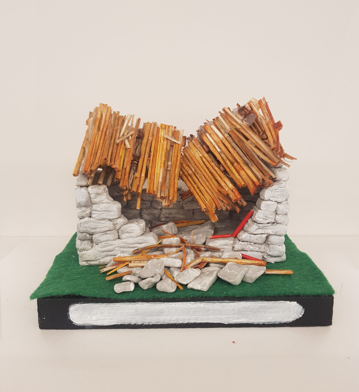 A photograph of an artwork representing a tiny replica of a ruined Irish stone cottage. The piece sits atop a square green felt, on a black base, similar to a traditional trophy stand. White paint at the front of the base indicates where a name might be, but there is no information given to identify the cottage. The cottage’s stones are made from a modelling material and painted grey. The front wall has mostly collapsed, with stones tumbling to rest on the green felt. Now the interior is visible through to the back wall. The roof, thatched with hollow orange-brown reeds, has also collapsed, sagging in the middle into the cottage’s interior. A few reeds have tumbled onto the pile of detached stones.