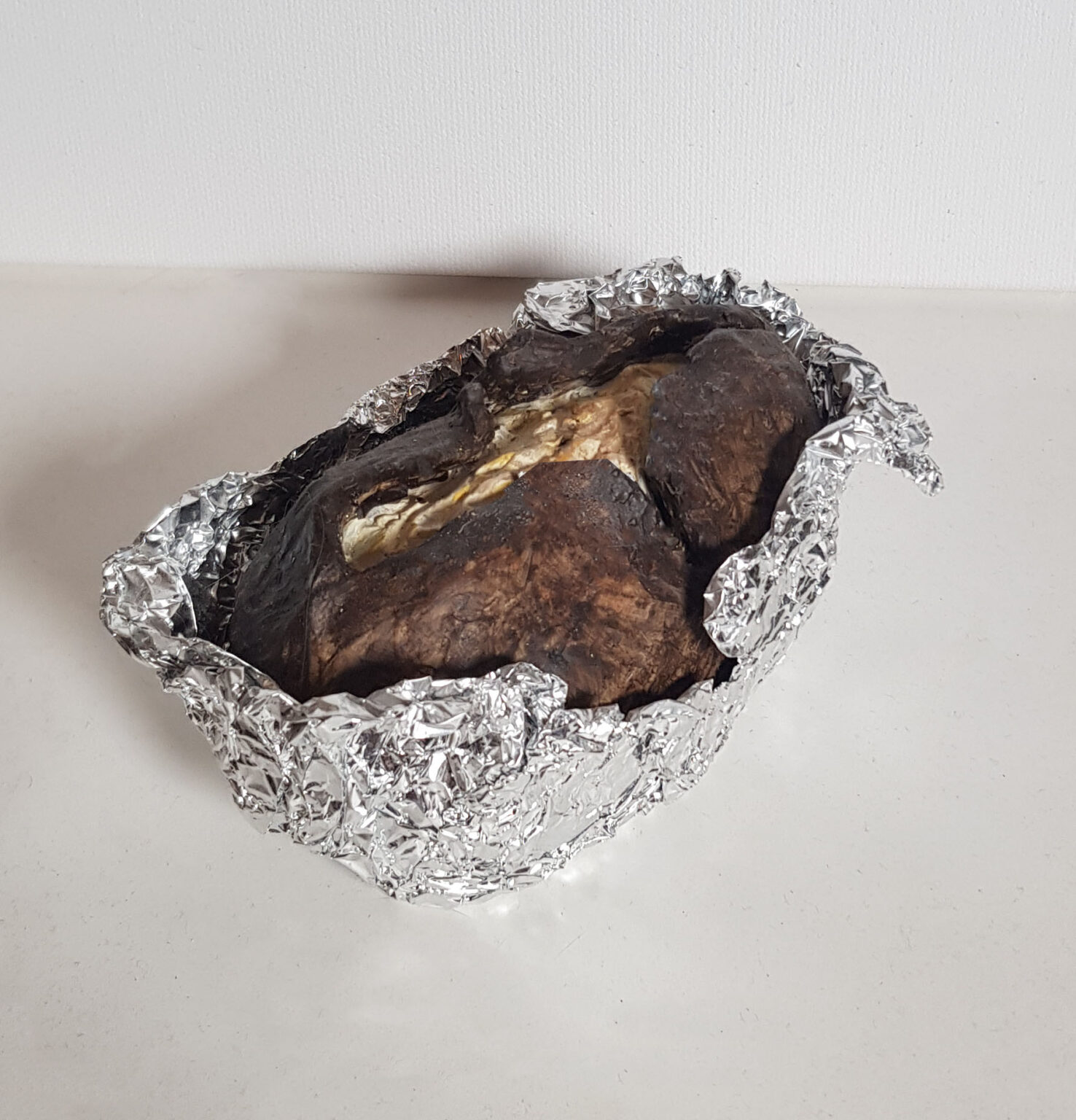 A photograph of a hyper-realistic sculpture of a baked potato. It sits within scrunched aluminium foil wrapping, open at the top. The potato’s skin, dark and almost burnt, is cut open in a cross shape, revealing the potato’s cooked flesh inside.