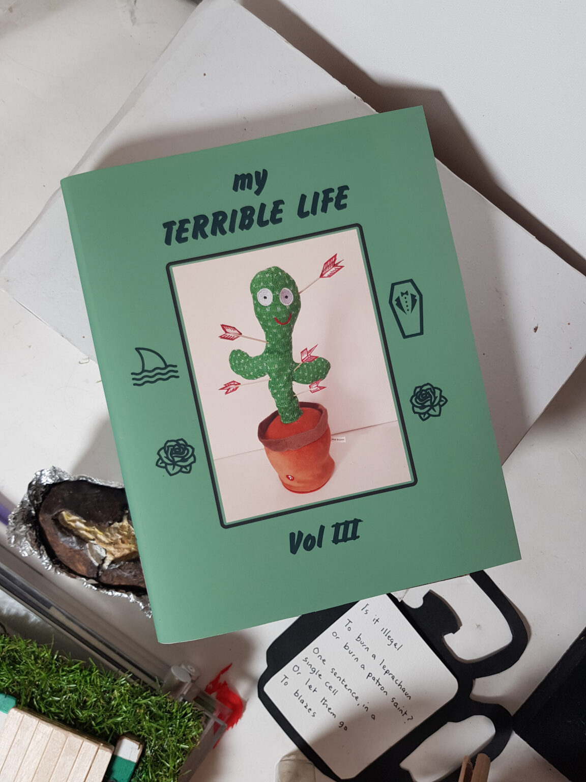 A photograph of an artwork that simulates a book cover. On green card, the title, my TERRIBLE LIFE, Volume 3, is printed in black italic lettering. The central image is a photograph of a cactus toy sitting in a flowerpot. The cactus is knitted, made from green wool with flecks of white. Red arrows sticking out of the cactus, nonetheless it has a big red smile and large inquisitive eye embroidered on as a face. A small label attached to the flower pot reads Made in China. Printed on the green cover around this image are four drawings in simple black line: a shark fin in water, a rose, a suit in a coffin and another rose. The book rests on a messy desk with a baked potato, fake grass and handwritten notes beginning with the Is it illegal.